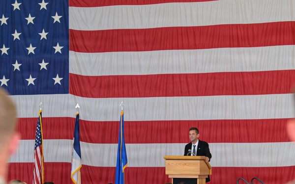 A civilian speaker in a dark suit addresses a large audience of military personnel seated in folding chairs at what appears to be an official ceremony or briefing. The event is held in a large indoor venue, likely a hangar or gymnasium, decorated with an oversized American flag as a backdrop. Three flags are displayed on poles behind the podium, including the U.S. flag and what appear to be Air Force-related flags.