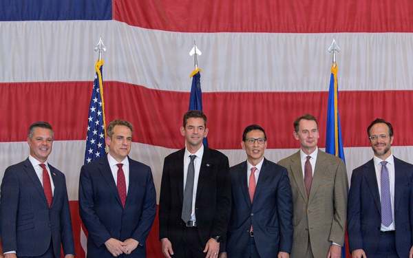 A formal group photo of eight men posing in front of a large American flag backdrop, along with what appears to be a U.S. Air Force flag. Two individuals on the far left and far right are wearing military flight suits, while the remaining six are dressed in business suits. The setting appears to be inside a military hangar or large facility, suggesting an official ceremony or announcement event.
