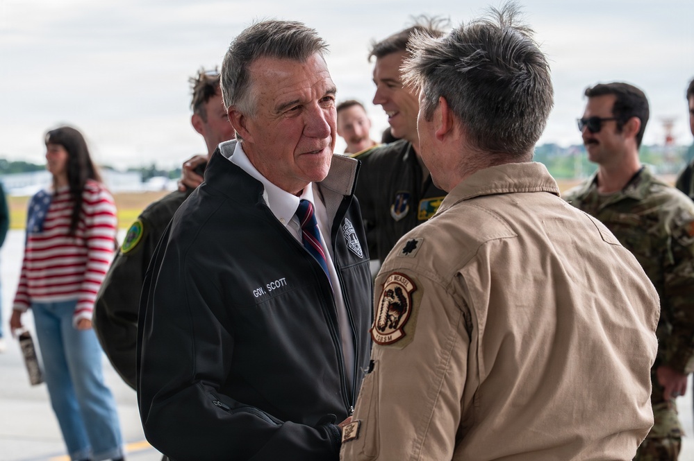 A person identified by their jacket as 'Gov. Scott' is shaking hands with a military officer in a tan flight suit on what appears to be an airfield tarmac. The setting suggests an official visit or ceremony at a military air installation. Several other individuals, including military personnel in flight suits and camouflage uniforms, are visible in the background.