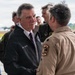 A person identified by their jacket as 'Gov. Scott' is shaking hands with a military officer in a tan flight suit on what appears to be an airfield tarmac. The setting suggests an official visit or ceremony at a military air installation. Several other individuals, including military personnel in flight suits and camouflage uniforms, are visible in the background.