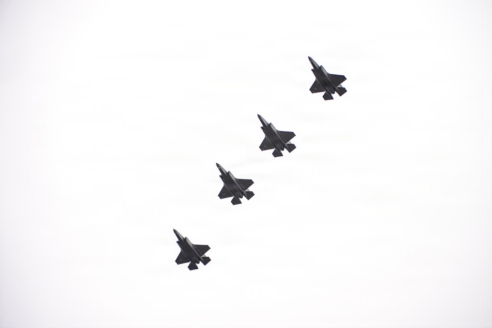 Four military jet aircraft are flying in a diagonal echelon formation against an overcast, white sky. The aircraft appear to be fifth-generation stealth fighters based on their visible silhouette characteristics, including delta wings and twin tail fins. The formation is evenly spaced and traveling in the same direction.
