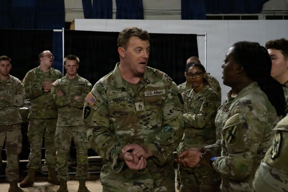 A group of U.S. Army soldiers in OCP (Operational Camouflage Pattern) uniforms are gathered in what appears to be an indoor facility, likely a gymnasium or assembly hall. The central figure, identified by his name tape as 'SADLER,' appears to be speaking or briefing a smaller group of soldiers in front of him. The setting suggests a unit meeting, training session, or pre-deployment briefing.