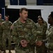 A group of U.S. Army soldiers in OCP (Operational Camouflage Pattern) uniforms are gathered in what appears to be an indoor facility, likely a gymnasium or assembly hall. The central figure, identified by his name tape as 'SADLER,' appears to be speaking or briefing a smaller group of soldiers in front of him. The setting suggests a unit meeting, training session, or pre-deployment briefing.