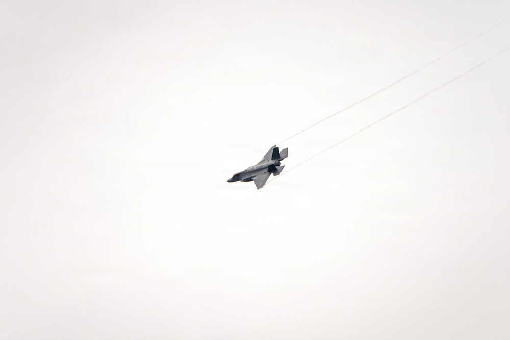 A single military jet aircraft is captured in flight against a pale, overcast sky. The aircraft appears to be a fifth-generation stealth fighter, consistent with an F-35 Lightning II, banking at an angle with visible vapor trails streaming from its wingtips. The image was taken from a ground-level perspective looking upward.