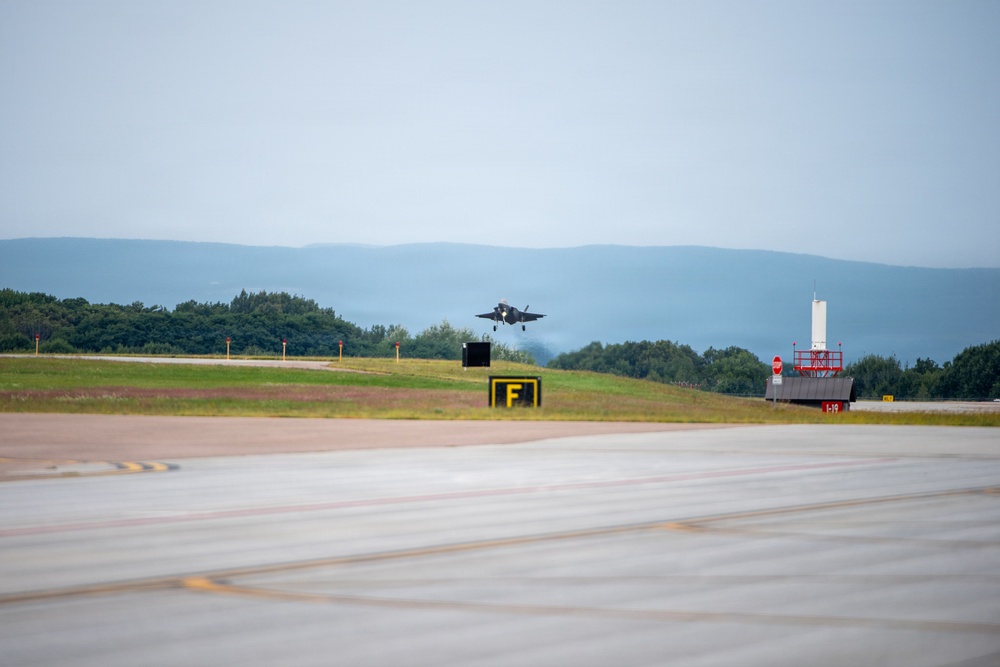 A military jet aircraft, consistent with an F-35 stealth fighter, is captured on final approach or low-altitude flight over a runway. The airfield appears to be a military or dual-use installation surrounded by forested hills and mountains in the background. Ground infrastructure including taxiway signage and approach lighting structures are visible.
