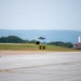 A military jet aircraft, consistent with an F-35 stealth fighter, is captured on final approach or low-altitude flight over a runway. The airfield appears to be a military or dual-use installation surrounded by forested hills and mountains in the background. Ground infrastructure including taxiway signage and approach lighting structures are visible.
