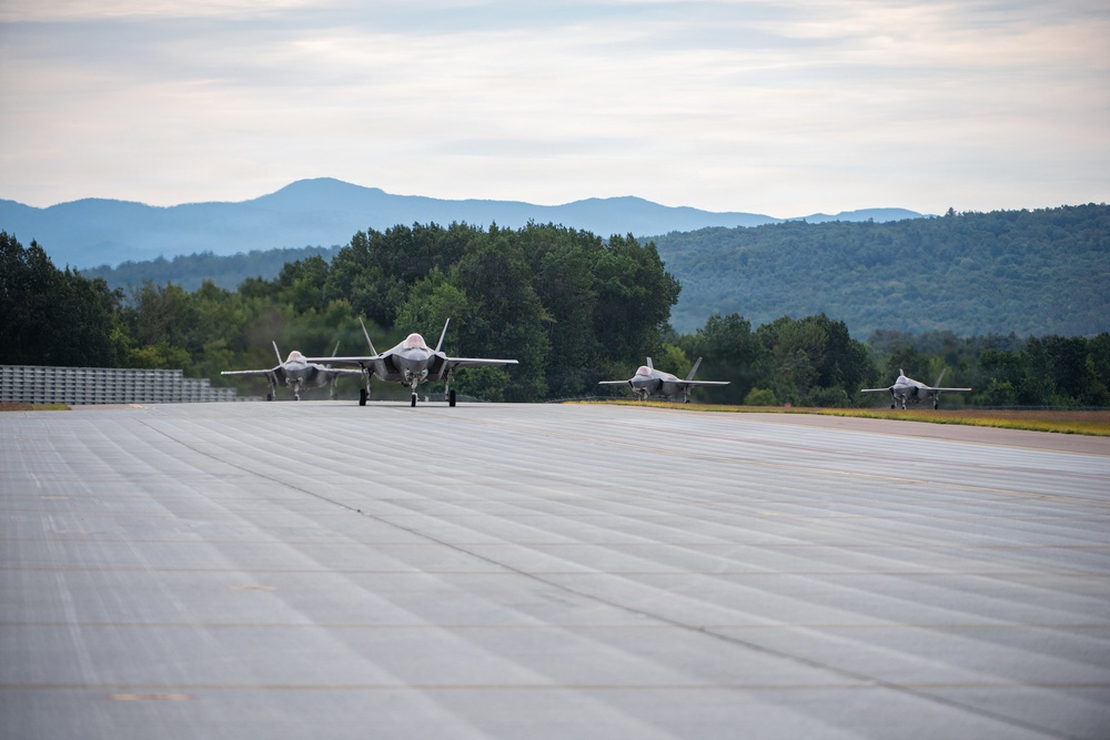 Four fifth-generation stealth fighter jets are taxiing on a runway in a two-by-two formation. The aircraft appear to be F-35 Lightning II jets based on their distinctive delta-wing, single-engine stealth design. The setting is a military airfield surrounded by dense green trees and a mountain range visible in the background under an overcast sky.