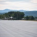 Four fifth-generation stealth fighter jets are taxiing on a runway in a two-by-two formation. The aircraft appear to be F-35 Lightning II jets based on their distinctive delta-wing, single-engine stealth design. The setting is a military airfield surrounded by dense green trees and a mountain range visible in the background under an overcast sky.