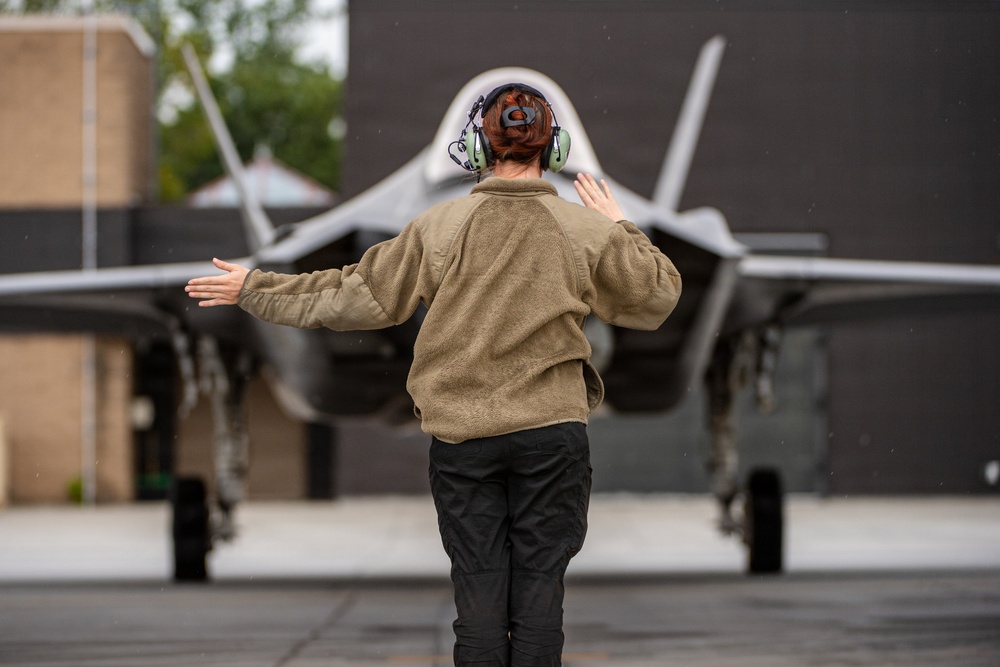 A military ground crew member is marshalling a fifth-generation stealth fighter jet on a tarmac. The person, viewed from behind, is using hand signals with arms outstretched to guide the aircraft. The scene appears to be taking place at a military airbase under overcast, possibly rainy conditions.