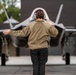A military ground crew member is marshalling a fifth-generation stealth fighter jet on a tarmac. The person, viewed from behind, is using hand signals with arms outstretched to guide the aircraft. The scene appears to be taking place at a military airbase under overcast, possibly rainy conditions.