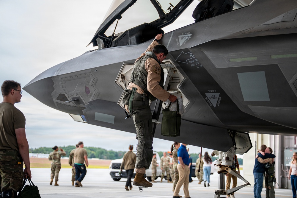 A military pilot in full flight gear is climbing down from or inspecting an F-35 Lightning II stealth fighter jet on a flight line tarmac. In the background, numerous military personnel and civilians are gathered, with some appearing to embrace in what suggests a homecoming or deployment return event. The setting appears to be a military air base with hangars visible in the background.