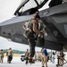 A military pilot in full flight gear is climbing down from or inspecting an F-35 Lightning II stealth fighter jet on a flight line tarmac. In the background, numerous military personnel and civilians are gathered, with some appearing to embrace in what suggests a homecoming or deployment return event. The setting appears to be a military air base with hangars visible in the background.