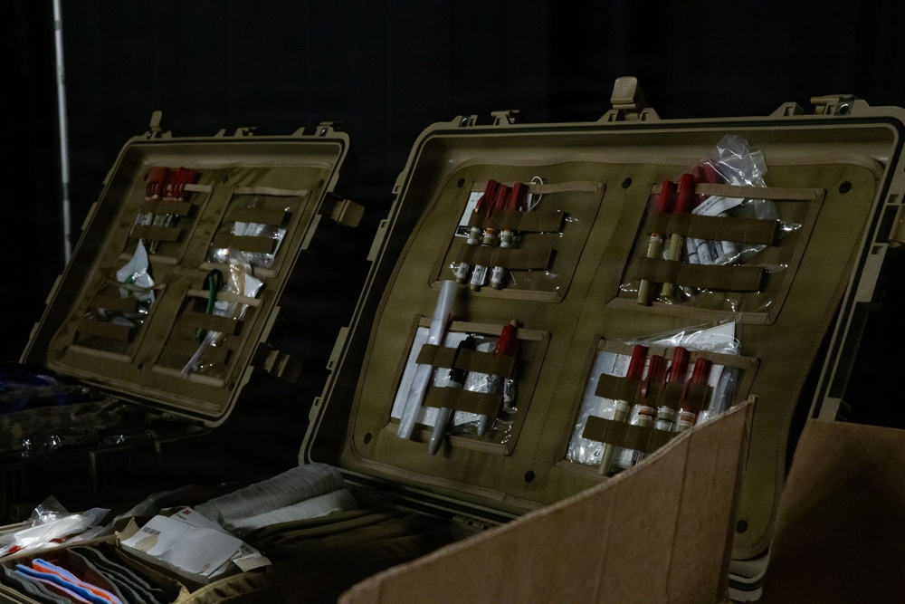 The image shows two open military-style hard-shell cases (likely Pelican-type cases) in olive drab/tan color, organized with medical supplies secured in MOLLE-compatible panel organizers on the lids. The cases contain what appear to be vials, syringes, packaged medical items, and other pharmaceutical or field medical supplies. The setting appears to be a dimly lit staging or operations area.