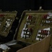 The image shows two open military-style hard-shell cases (likely Pelican-type cases) in olive drab/tan color, organized with medical supplies secured in MOLLE-compatible panel organizers on the lids. The cases contain what appear to be vials, syringes, packaged medical items, and other pharmaceutical or field medical supplies. The setting appears to be a dimly lit staging or operations area.