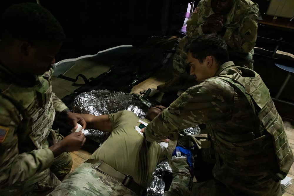 Military personnel in camouflage uniforms are conducting a tactical combat casualty care (TCCC) training exercise. Two soldiers are treating a simulated casualty lying on the floor wrapped in a mylar emergency blanket, with one soldier applying bandaging to the casualty's arm. The scene appears to be an indoor training environment with low lighting conditions.