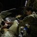 Military personnel in camouflage uniforms are conducting a tactical combat casualty care (TCCC) training exercise. Two soldiers are treating a simulated casualty lying on the floor wrapped in a mylar emergency blanket, with one soldier applying bandaging to the casualty's arm. The scene appears to be an indoor training environment with low lighting conditions.