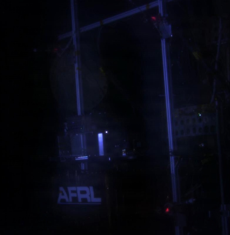A very dark image showing what appears to be a laboratory or technical facility setup with structural framework visible. The acronym 'AFRL' (Air Force Research Laboratory) is prominently illuminated in white text in the lower portion of the image. Small colored indicator lights (red and green) are visible on various pieces of equipment within a metal frame structure.
