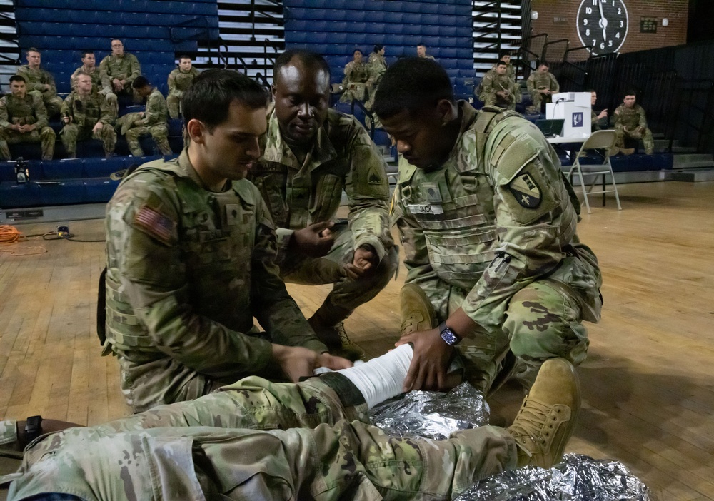 U.S. Army soldiers are conducting a combat casualty care (TCCC) training exercise inside a gymnasium or indoor arena. Three soldiers in OCP (Operational Camouflage Pattern) uniforms are actively practicing wound bandaging techniques on a simulated casualty lying on a mylar emergency blanket. A larger group of soldiers observes from the bleachers in the background, indicating a formal training event.