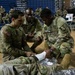 U.S. Army soldiers are conducting a combat casualty care (TCCC) training exercise inside a gymnasium or indoor arena. Three soldiers in OCP (Operational Camouflage Pattern) uniforms are actively practicing wound bandaging techniques on a simulated casualty lying on a mylar emergency blanket. A larger group of soldiers observes from the bleachers in the background, indicating a formal training event.