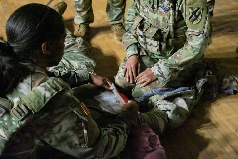 Two U.S. Army soldiers in Operational Camouflage Pattern (OCP) uniforms are engaged in what appears to be a combat first aid or medical training exercise on a gymnasium floor. One soldier is applying or demonstrating the use of a bandage or medical dressing on another soldier's leg area. The setting appears to be an indoor training facility with a hardwood floor.