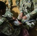 Two U.S. Army soldiers in Operational Camouflage Pattern (OCP) uniforms are engaged in what appears to be a combat first aid or medical training exercise on a gymnasium floor. One soldier is applying or demonstrating the use of a bandage or medical dressing on another soldier's leg area. The setting appears to be an indoor training facility with a hardwood floor.