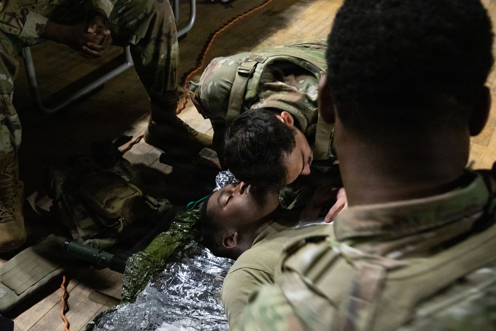 Military personnel in camouflage uniforms are conducting a combat casualty care or first aid training exercise. One soldier is performing a check on a simulated casualty lying on the floor wrapped in a mylar emergency blanket. Several other soldiers in OCP (Operational Camouflage Pattern) uniforms are observing the procedure.