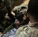 Military personnel in camouflage uniforms are conducting a combat casualty care or first aid training exercise. One soldier is performing a check on a simulated casualty lying on the floor wrapped in a mylar emergency blanket. Several other soldiers in OCP (Operational Camouflage Pattern) uniforms are observing the procedure.