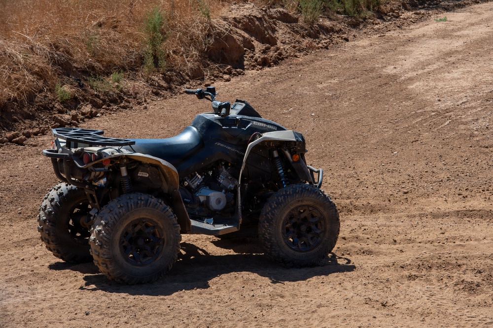 A black Can-Am Renegade ATV is parked on a dry, sandy dirt trail in an arid outdoor environment. The vehicle appears to be a high-performance off-road quad bike with knobby tires and visible engine components. The surrounding terrain features dry brush, reddish-brown soil, and eroded embankments typical of a desert or semi-arid region.