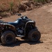 A black Can-Am Renegade ATV is parked on a dry, sandy dirt trail in an arid outdoor environment. The vehicle appears to be a high-performance off-road quad bike with knobby tires and visible engine components. The surrounding terrain features dry brush, reddish-brown soil, and eroded embankments typical of a desert or semi-arid region.