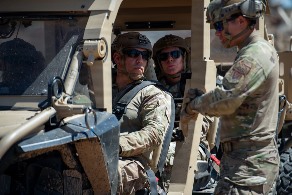 Three military personnel in full combat gear are positioned in and around a tan/desert-colored tactical military vehicle. Two soldiers are seated inside the open-frame vehicle while a third stands outside engaging with them. All personnel are wearing MultiCam (OCP) uniforms, tactical helmets, and ballistic sunglasses.