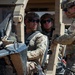Three military personnel in full combat gear are positioned in and around a tan/desert-colored tactical military vehicle. Two soldiers are seated inside the open-frame vehicle while a third stands outside engaging with them. All personnel are wearing MultiCam (OCP) uniforms, tactical helmets, and ballistic sunglasses.