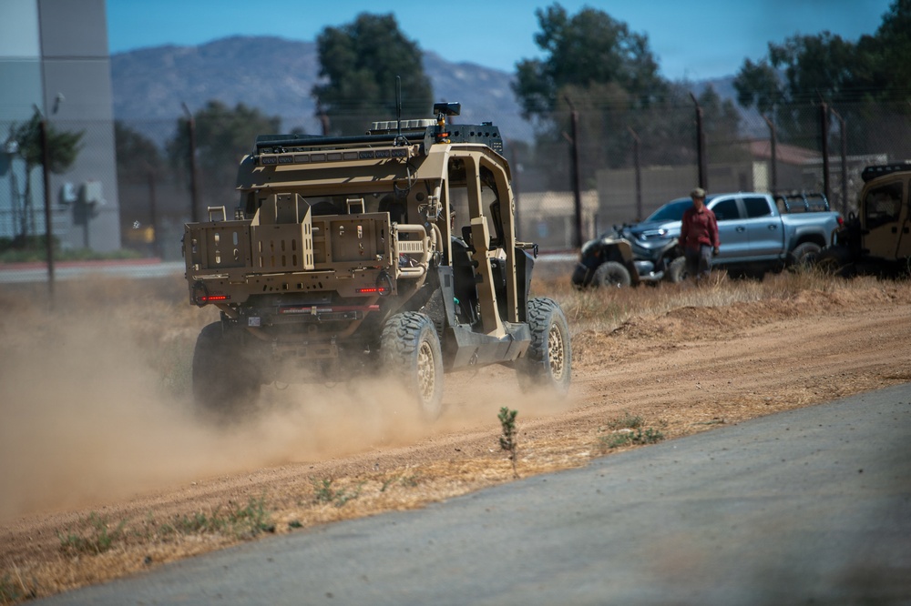 A tan/desert-colored military tactical utility vehicle (UTV) is driving along a dirt road, kicking up a dust cloud. The scene appears to be a military base or training facility, with additional vehicles and a person visible in the background. Mountains and trees are visible in the distance under a clear blue sky.