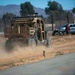 A tan/desert-colored military tactical utility vehicle (UTV) is driving along a dirt road, kicking up a dust cloud. The scene appears to be a military base or training facility, with additional vehicles and a person visible in the background. Mountains and trees are visible in the distance under a clear blue sky.