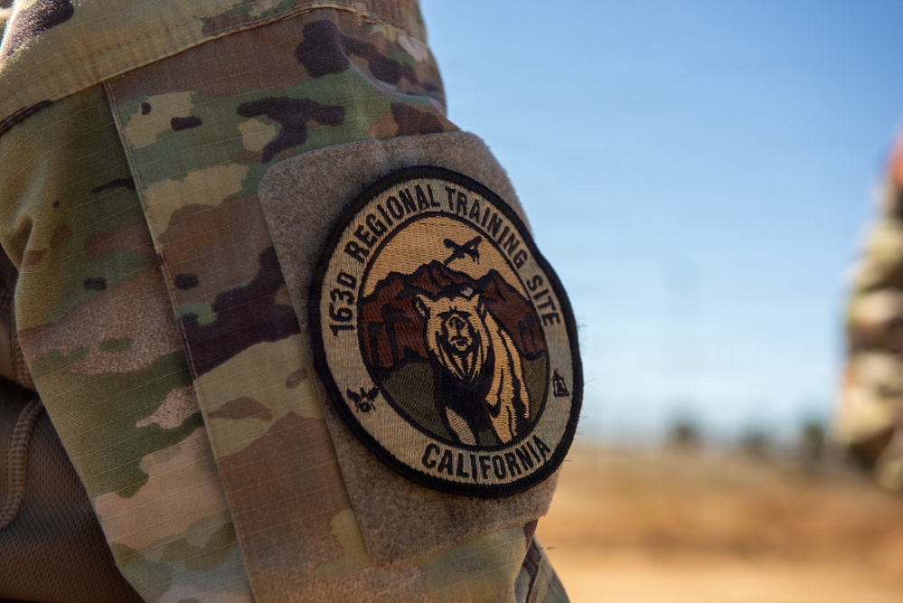 A close-up photograph of a military uniform sleeve displaying a circular embroidered patch for the 163d Regional Training Site, California. The patch features a grizzly bear against a mountain backdrop with aircraft silhouettes. The uniform is MultiCam/OCP camouflage pattern, and the patch is attached via a hook-and-loop (Velcro) field.