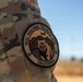 A close-up photograph of a military uniform sleeve displaying a circular embroidered patch for the 163d Regional Training Site, California. The patch features a grizzly bear against a mountain backdrop with aircraft silhouettes. The uniform is MultiCam/OCP camouflage pattern, and the patch is attached via a hook-and-loop (Velcro) field.