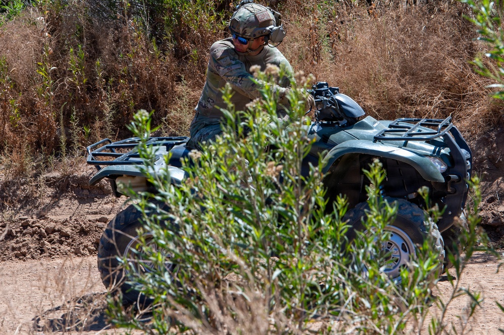 A person in military camouflage uniform and tactical helmet is operating an all-terrain vehicle (ATV) on a dirt trail through dry, brushy terrain. The rider is leaning forward on the quad bike, navigating through sparse vegetation and sandy soil. The setting appears to be a rural or training area with dry grass and shrubs in the background.