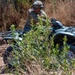 A person in military camouflage uniform and tactical helmet is operating an all-terrain vehicle (ATV) on a dirt trail through dry, brushy terrain. The rider is leaning forward on the quad bike, navigating through sparse vegetation and sandy soil. The setting appears to be a rural or training area with dry grass and shrubs in the background.
