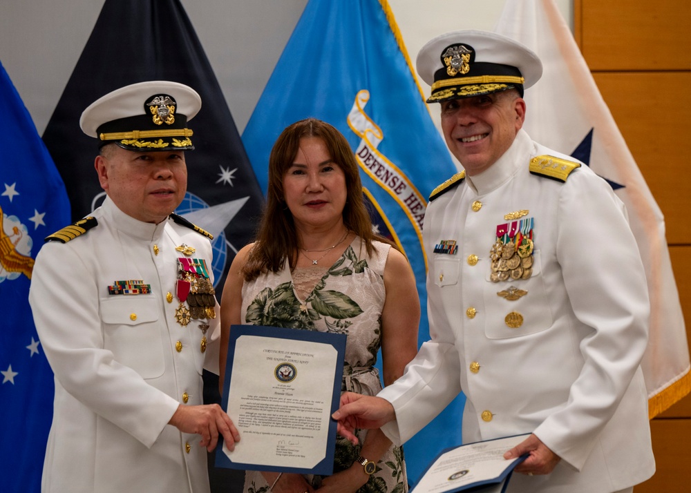 Two U.S. Navy officers in white dress uniforms flank a civilian woman during what appears to be a formal military ceremony. The group is posing together while holding a blue-covered certificate or commendation document. The setting includes multiple military flags in the background, including a visible 'Defense Health' flag.