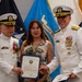 Two U.S. Navy officers in white dress uniforms flank a civilian woman during what appears to be a formal military ceremony. The group is posing together while holding a blue-covered certificate or commendation document. The setting includes multiple military flags in the background, including a visible 'Defense Health' flag.