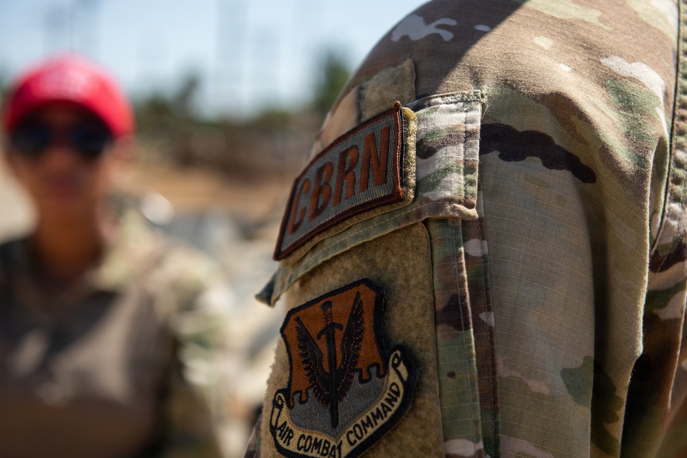 A close-up photograph of a U.S. Air Force service member's left sleeve, showing two distinct patches on an OCP (Operational Camouflage Pattern) uniform. The patches identify the wearer as belonging to Air Combat Command and specializing in CBRN (Chemical, Biological, Radiological, and Nuclear) defense. A second individual wearing a red cap and sunglasses is visible but blurred in the background.