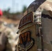 A close-up photograph of a U.S. Air Force service member's left sleeve, showing two distinct patches on an OCP (Operational Camouflage Pattern) uniform. The patches identify the wearer as belonging to Air Combat Command and specializing in CBRN (Chemical, Biological, Radiological, and Nuclear) defense. A second individual wearing a red cap and sunglasses is visible but blurred in the background.