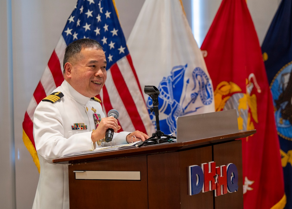 A military officer in a white dress uniform is speaking at a podium labeled 'DHHQ' while holding a microphone. He is smiling and appears to be delivering a speech or presentation, with a laptop visible on the podium. Multiple flags, including the United States flag, a Marine Corps flag, and other military/government flags, are displayed in the background.