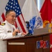 A military officer in a white dress uniform is speaking at a podium labeled 'DHHQ' while holding a microphone. He is smiling and appears to be delivering a speech or presentation, with a laptop visible on the podium. Multiple flags, including the United States flag, a Marine Corps flag, and other military/government flags, are displayed in the background.