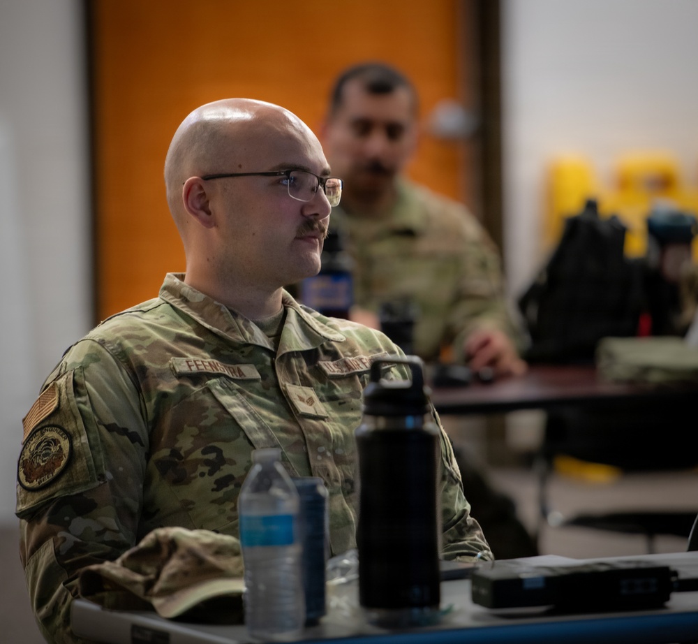 A U.S. Air Force service member wearing OCP (Operational Camouflage Pattern) uniform with the name tape 'FEENSTRA' is seated at a table, appearing to attend a briefing or training session. A second service member is visible in the background, also in OCP uniform. The foreground subject wears glasses and has a shaved head with a mustache.