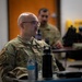 A U.S. Air Force service member wearing OCP (Operational Camouflage Pattern) uniform with the name tape 'FEENSTRA' is seated at a table, appearing to attend a briefing or training session. A second service member is visible in the background, also in OCP uniform. The foreground subject wears glasses and has a shaved head with a mustache.