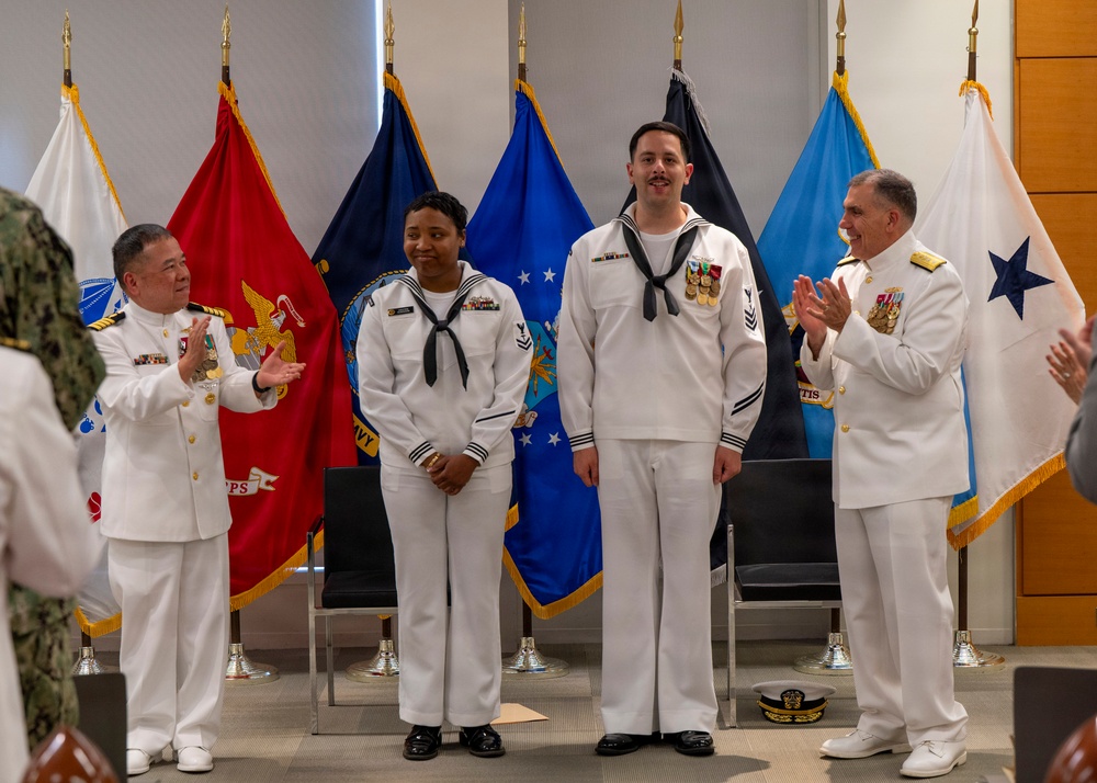 A U.S. Navy military ceremony is taking place in an indoor setting, likely a promotion or award ceremony. Four personnel in white dress uniforms are standing before a display of military service branch flags, with two senior officers applauding two junior enlisted sailors. The atmosphere appears celebratory and formal.