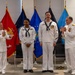 A U.S. Navy military ceremony is taking place in an indoor setting, likely a promotion or award ceremony. Four personnel in white dress uniforms are standing before a display of military service branch flags, with two senior officers applauding two junior enlisted sailors. The atmosphere appears celebratory and formal.
