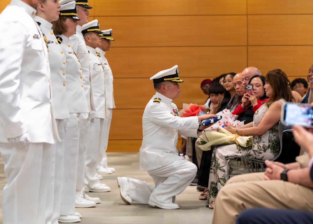 A U.S. Navy officer in dress white uniform is kneeling on one knee to present a folded American flag to a seated civilian woman, likely during a military retirement or memorial ceremony. A row of Navy personnel in dress whites stands at attention in the background. Audience members are photographing the moment with smartphones.