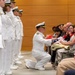 A U.S. Navy officer in dress white uniform is kneeling on one knee to present a folded American flag to a seated civilian woman, likely during a military retirement or memorial ceremony. A row of Navy personnel in dress whites stands at attention in the background. Audience members are photographing the moment with smartphones.