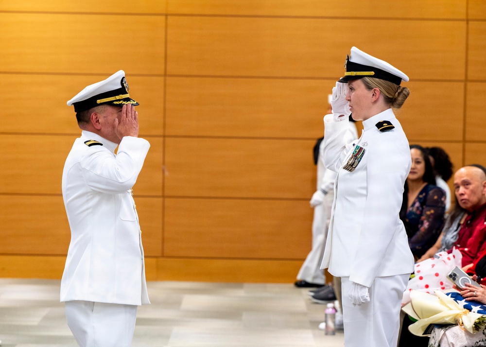 Two U.S. Navy officers in white dress uniforms are exchanging salutes during what appears to be a formal military ceremony. A male officer on the left and a female officer on the right face each other mid-salute, with the female officer displaying multiple service ribbons/medals on her uniform. Seated civilians and a third uniformed individual are visible in the background.